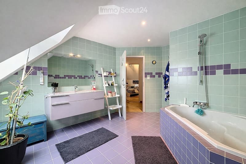 Bathroom with green and purple tiles, a bathtub, sink, mirror, and a small plant in a pot. The room features a slanted ceiling and an open doorway leading to another room.