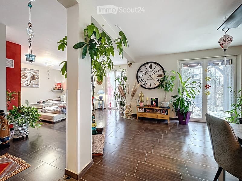 A spacious, well-lit room with wooden flooring, large plants, a sofa, a table, a chair, a clock, vases, a lamp, picture frames, and a bed.