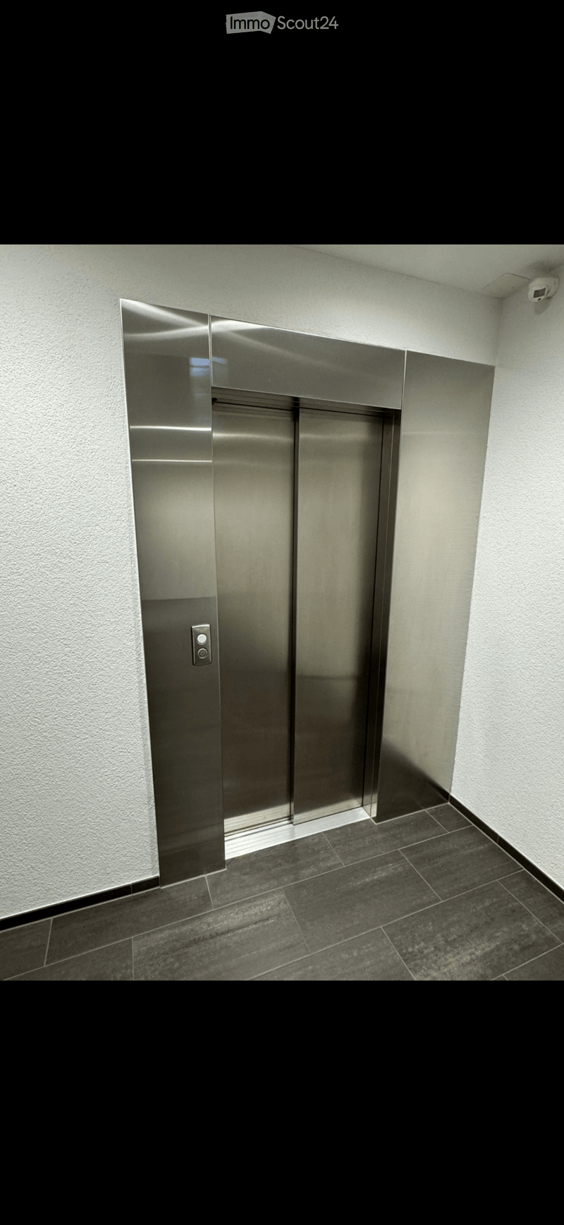 Modern hallway with dark tiled floor, light-colored walls, and a stainless steel elevator door with control panel.