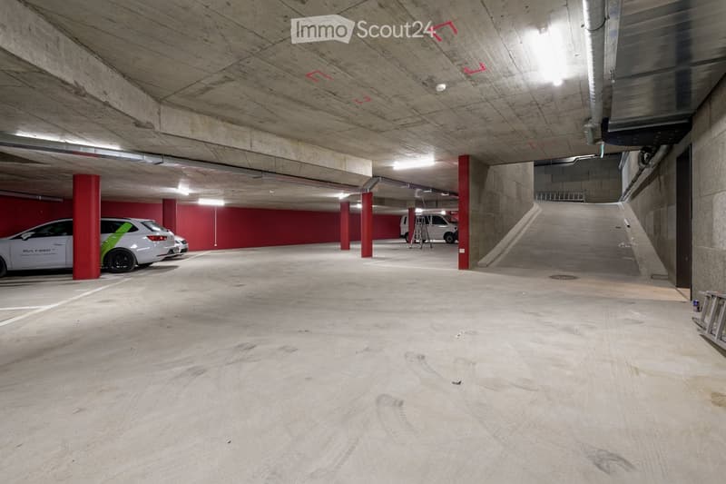 Underground parking area with concrete floors and walls, red support pillars, and several parked cars.