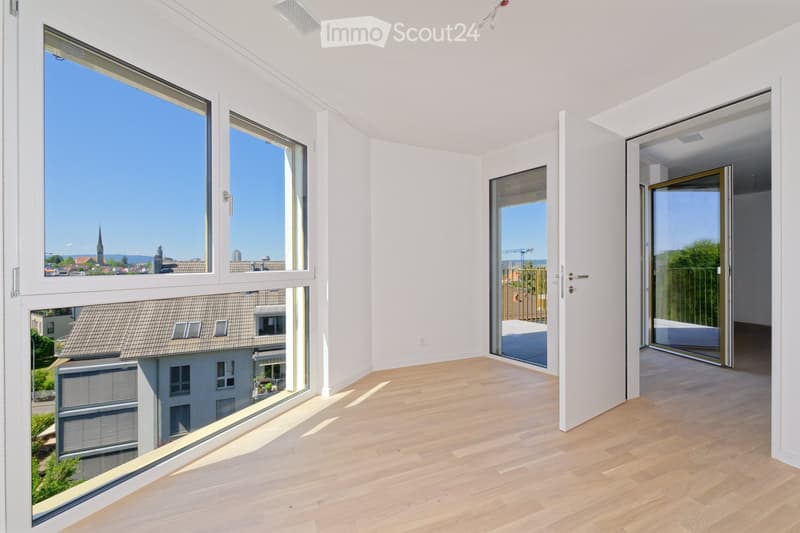 Bright, empty room with light wooden flooring, large windows, and sliding glass doors leading to a balcony or terrace. The view shows a cityscape with rooftops and a distant church spire.
