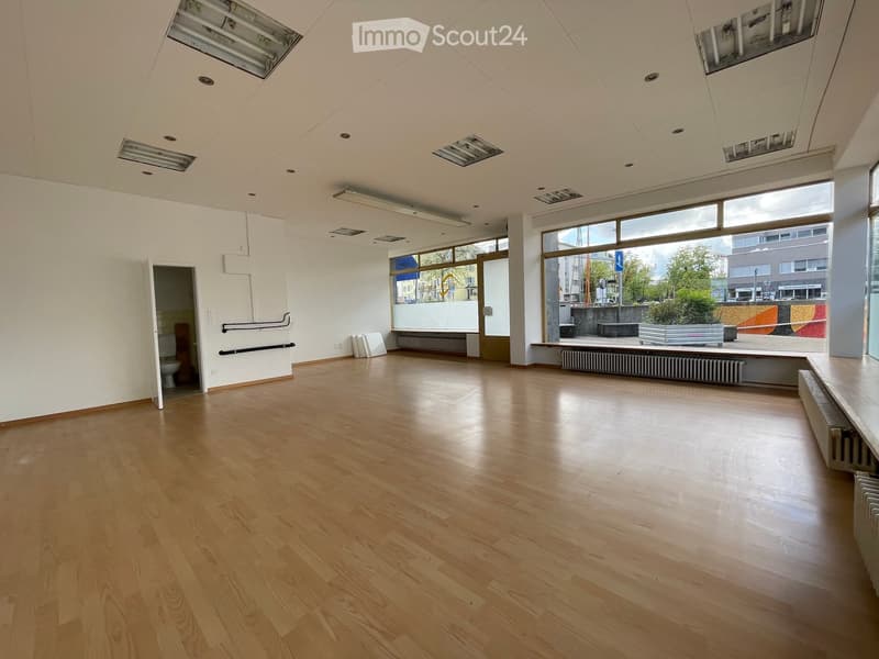 Empty room with wooden flooring, white walls, large windows, and a visible bathroom entrance.
