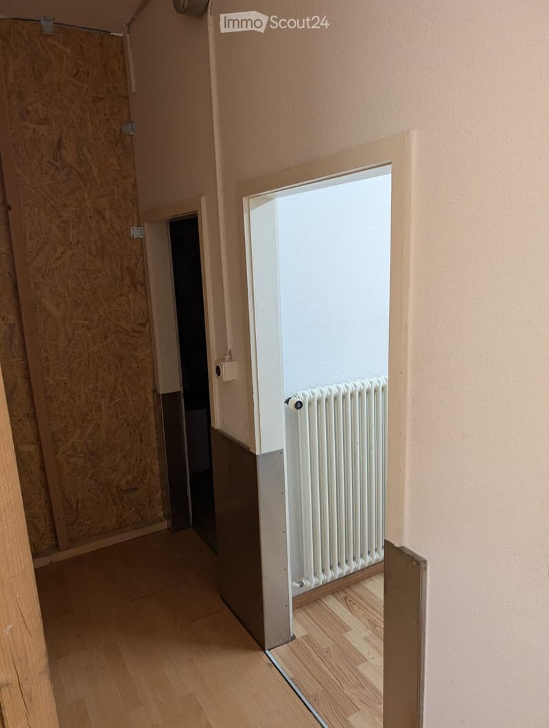 Empty hallway with light wooden flooring, beige walls, and a white radiator visible in an adjacent room.