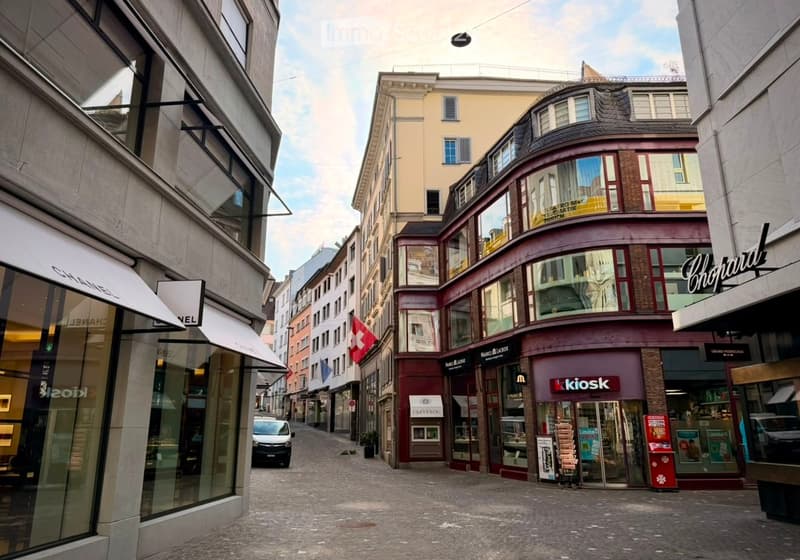 A narrow urban street lined with multi-story buildings, featuring shops with awnings and signage, including a visible Swiss flag.