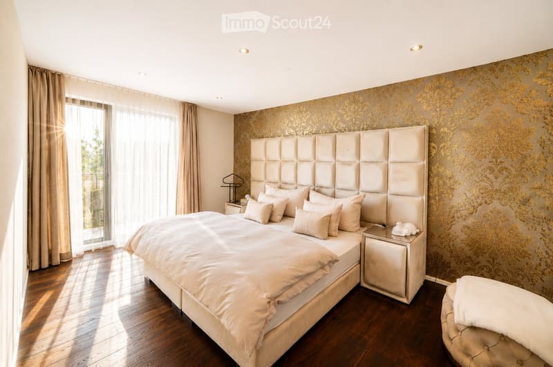 Elegant bedroom with a large upholstered headboard, a neatly made bed with multiple pillows, a nightstand with a lamp, and a small stool near the window.