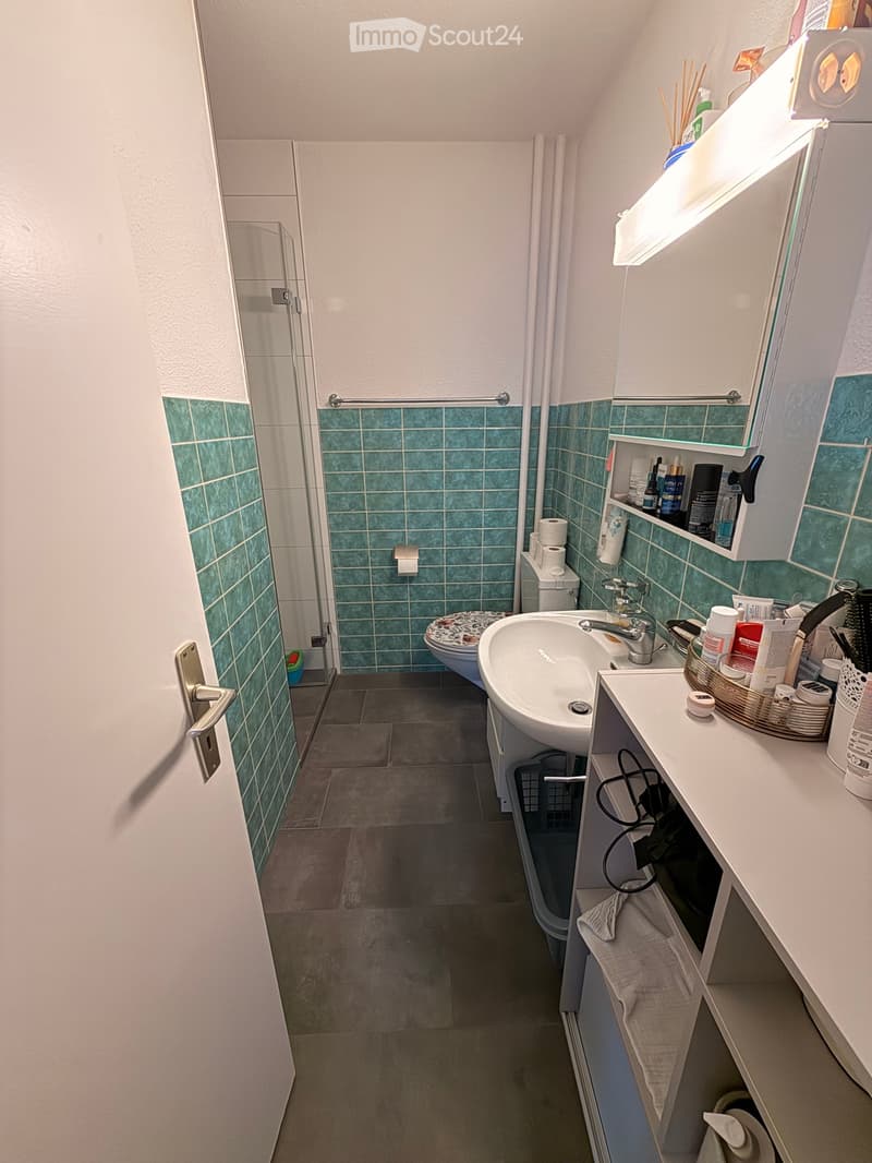 Bathroom with green tiled walls, a sink with a mirror cabinet, a toilet, and a shower area with a glass door. The floor is tiled, and there is a towel on the cabinet.