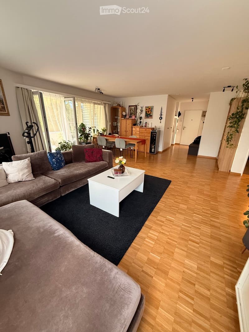 A modern living room with a grey sectional sofa, white coffee table, dining area with a wooden table and chairs, and a black exercise bike in the corner. The room features wooden flooring, large windows with curtains, and several decorative items includin