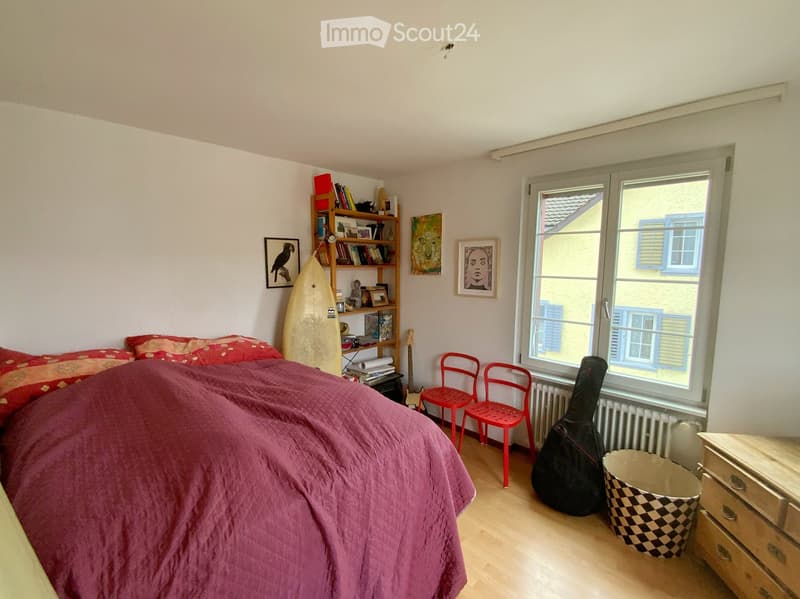 Cozy bedroom with a double bed covered in a purple blanket, red chairs, a guitar and case, a bookshelf with books and a surfboard, and framed pictures on the walls.