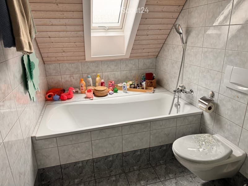 Modern bathroom with a bathtub, toilet, and showerhead. The room features marble tiles, a skylight window, and various toiletries and accessories on the edge of the bathtub.