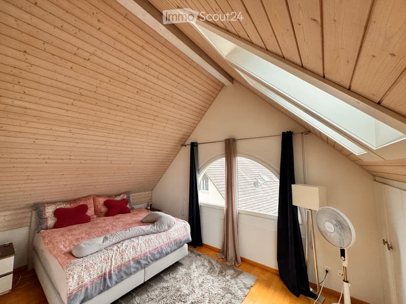 Cozy attic bedroom with wooden slanted ceilings, skylights, and large windows. Features a double bed with pink bedding, a nightstand, a standing fan, and a soft rug on the floor.