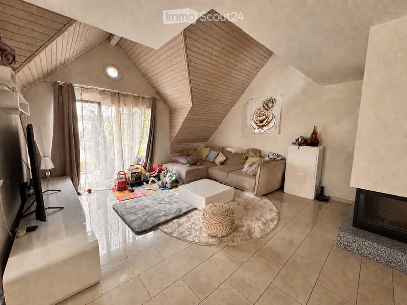 Bright living room with a slanted wooden ceiling, large window with curtains, beige sofa, coffee table, ottoman, floor lamp, television, fireplace, and decorative vases.