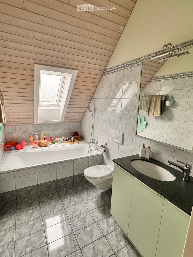 Bathroom with a bathtub, toilet, sink, mirror, and various toiletries on the windowsill. The room features wooden paneling on the ceiling and grey tiled walls and floor.