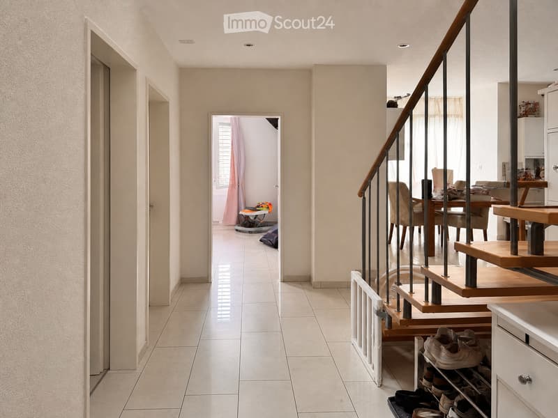 A well-lit hallway with tiled flooring, a staircase with wooden steps and metal railings, and a shoe rack with several pairs of shoes. The hallway leads to an open area with a dining table and chairs visible in the background.