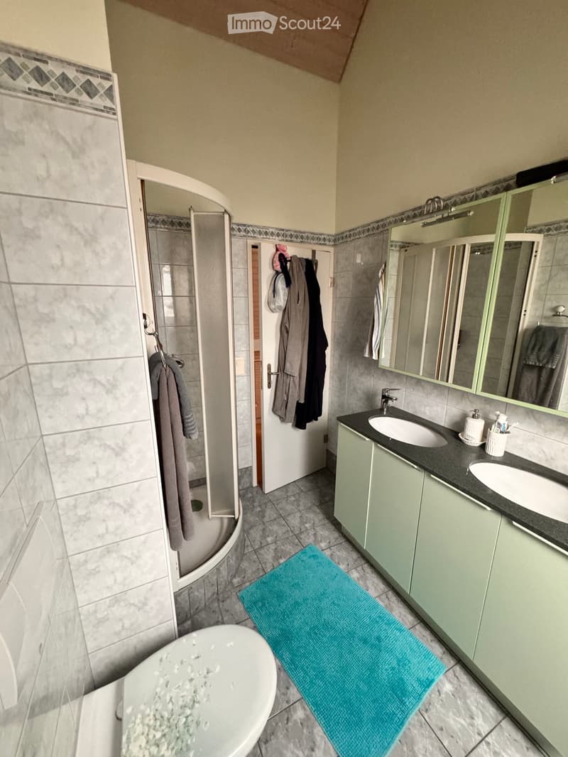 Modern bathroom with a bathtub, double sink, large mirror, and a shower area with a glass door. The space includes a blue bath mat, towels, and a cabinet with toiletries.