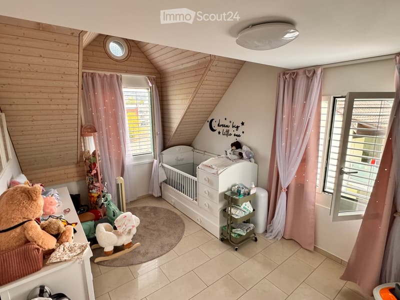 Cozy children's bedroom with wooden slanted ceilings, crib, rocking horse, toy cart, teddy bear, mirror, and pink curtains.
