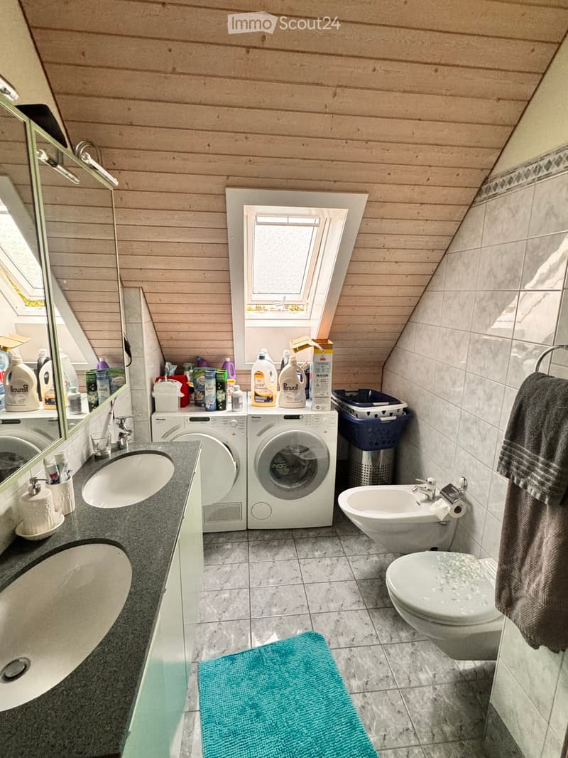 Bathroom with a sink, toilet, washing machine, and shower. The room features wooden paneling on the ceiling and walls, with a window providing natural light.