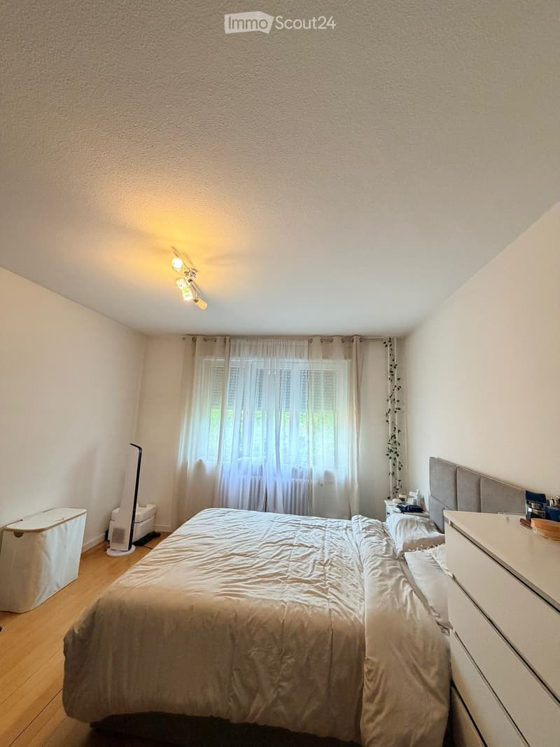 A well-lit bedroom with a large bed, nightstand, fan, and window with curtains.