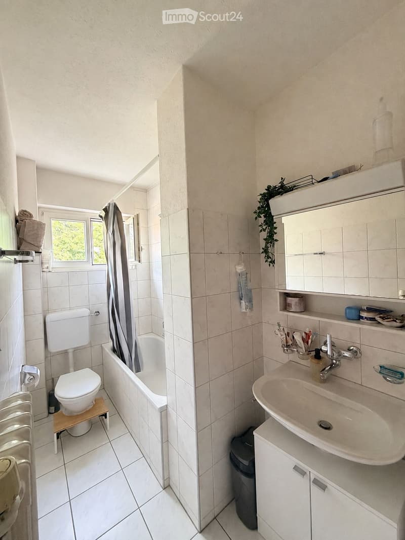 Bright bathroom with white tiles, a toilet, bathtub, sink, and mirror. Includes a small wooden stool under the toilet, a towel rack, and a small window with a view of greenery.