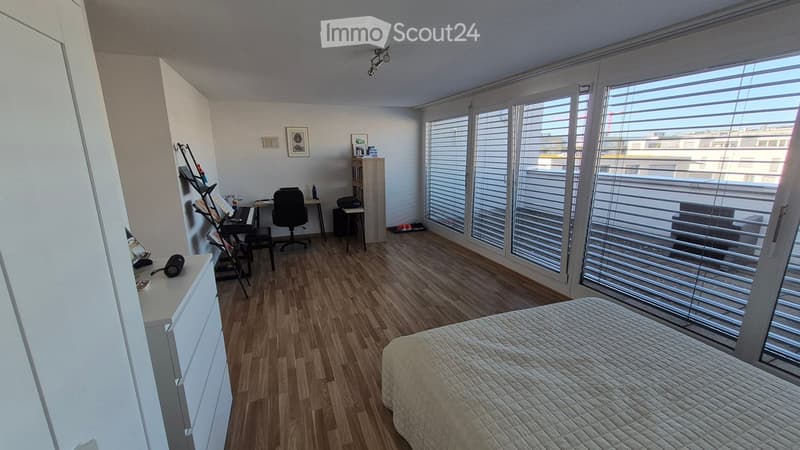 A modern, well-lit bedroom with a large window, wooden flooring, a neatly made bed, a desk with a chair, a bookshelf, and a lamp.