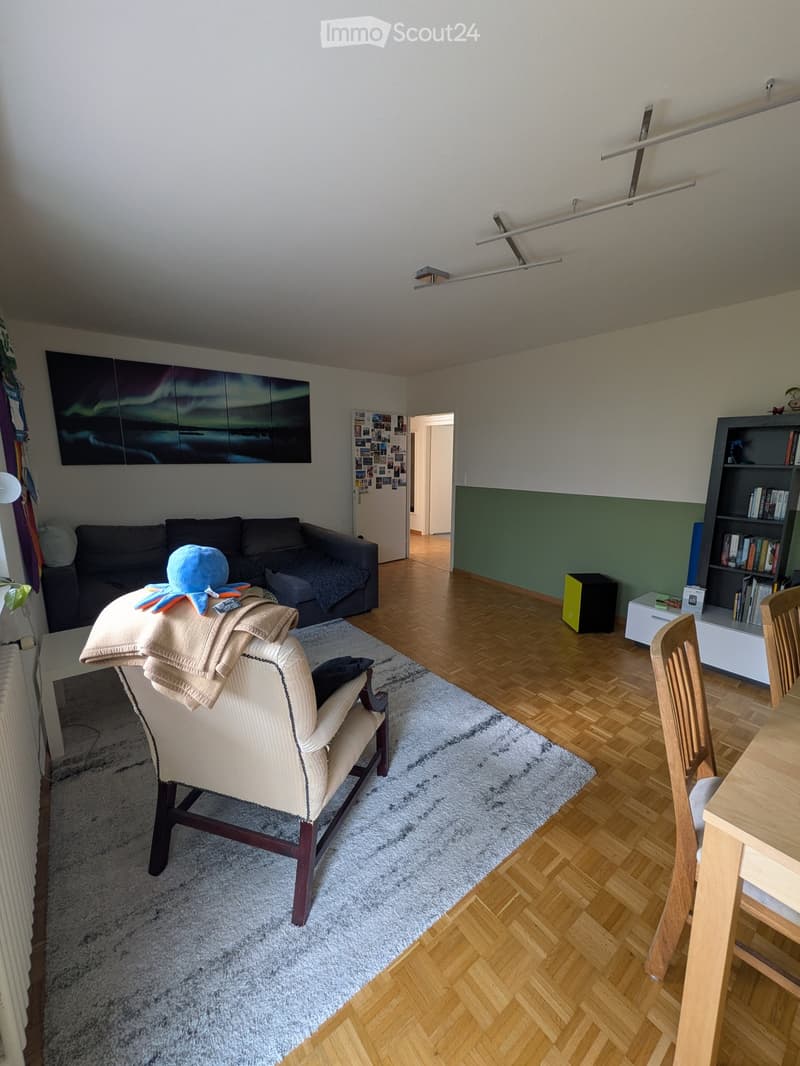 Living room with a sofa, armchair, coffee table, dining table, bookshelf, and a large painting on the wall.