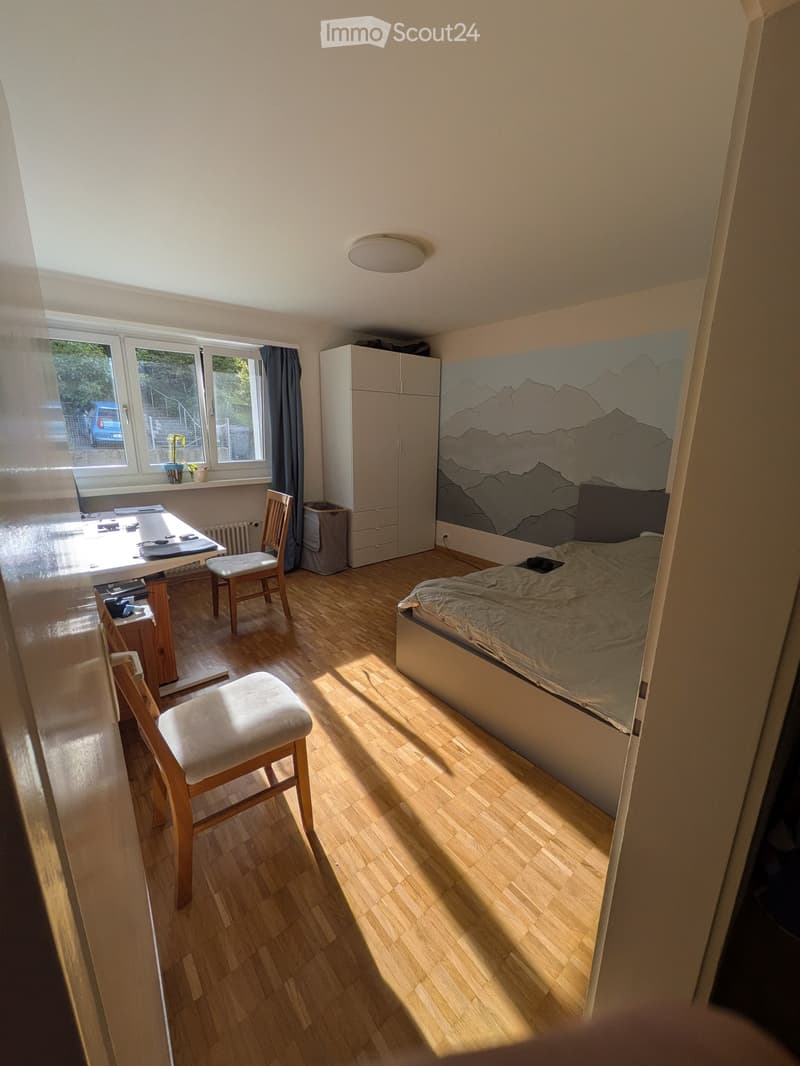 Bright bedroom with wooden flooring, a bed with a mountain-themed headboard, a small table with chairs, and a cabinet.