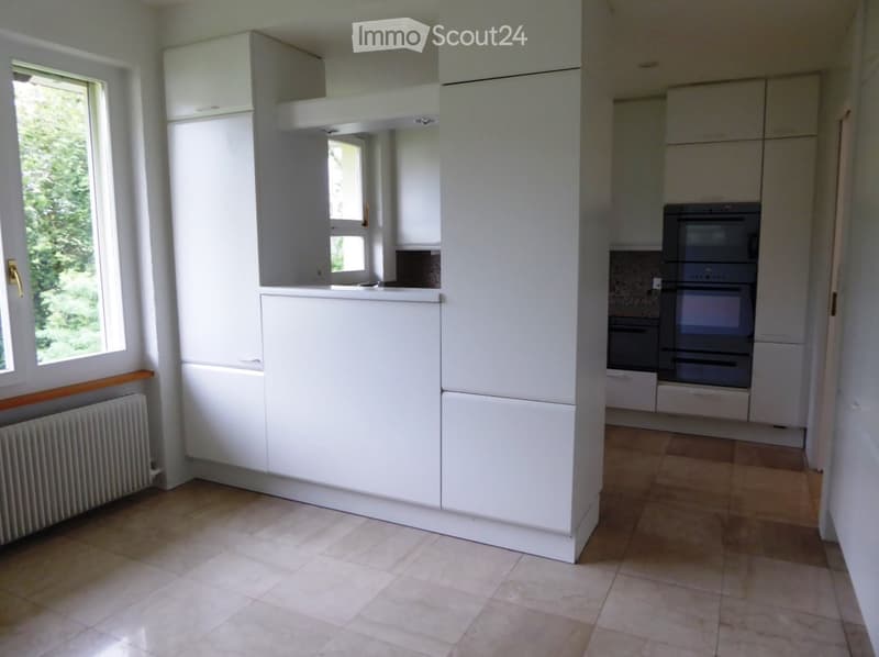 Modern kitchen with white cabinets, tiled floor, and built-in oven. Large windows provide natural light.