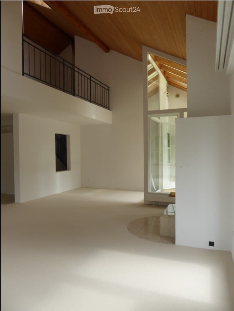 Empty room with white walls, wooden ceiling, and a balcony overlooking the space. The floor is light-colored and polished.