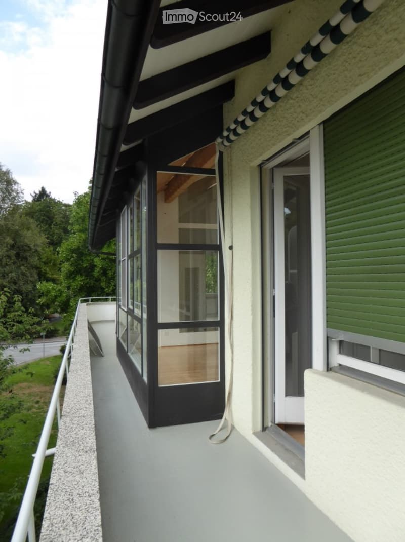Balcony with white railing, grey concrete floor, large windows with black frames, view of greenery and road.