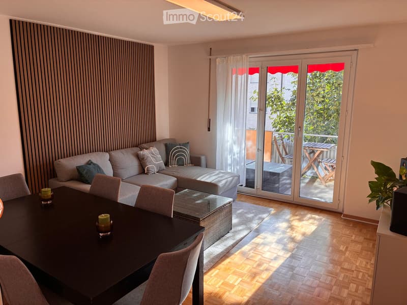 Bright living room with a grey sofa, ottoman, dining table, chairs, and a view to a balcony with outdoor furniture.
