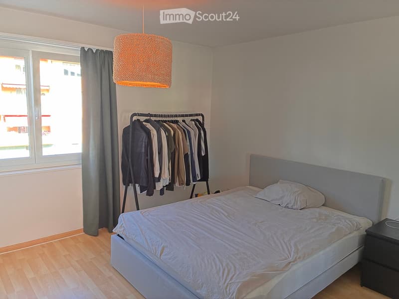 Cozy bedroom with a double bed, nightstand, hanging clothes, and a large window with a curtain.