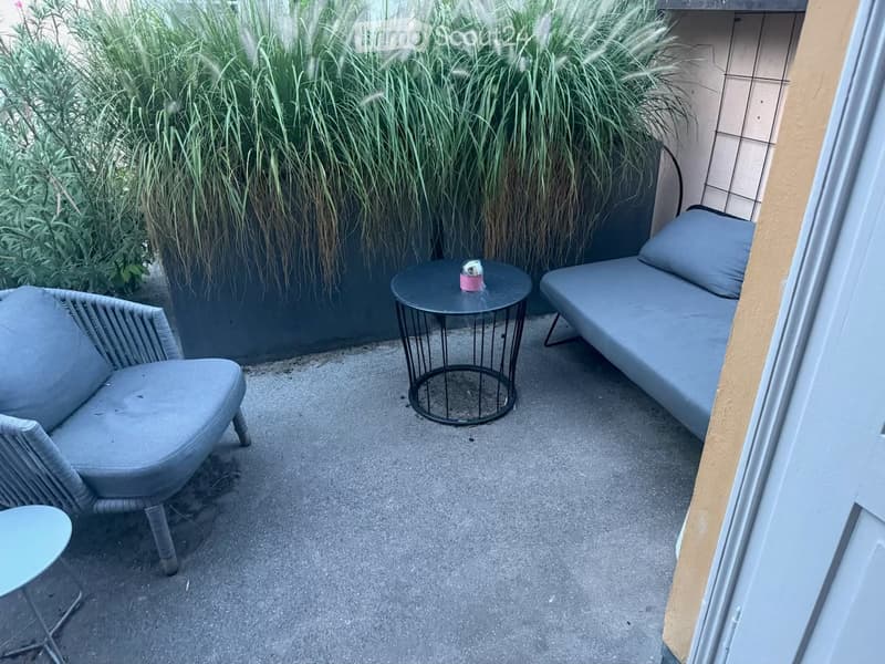 Balcony with a grey sofa, a small round table, and a wicker chair, surrounded by tall green plants.