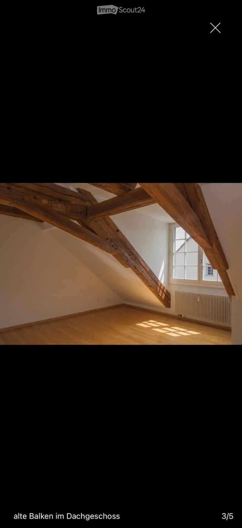 Empty attic room with wooden beams, wooden floor, large window, radiator, and exposed wooden beams on the ceiling.