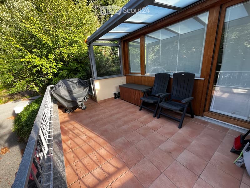 Balcony with tiled floor, two black chairs, a small table, and a covered grill, surrounded by greenery.