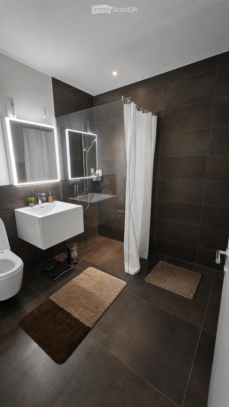 Modern bathroom with dark brown tiles, white sink, toilet, bathtub, and illuminated mirrors. Includes a shower area with a glass enclosure and a white shower curtain.