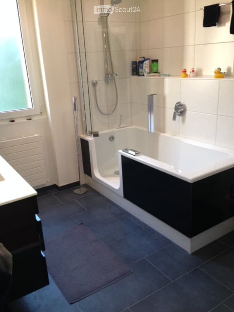 Modern bathroom with a white bathtub, shower area, sink, and tiled walls and floor. Includes a towel and small decorative items on the shelf.