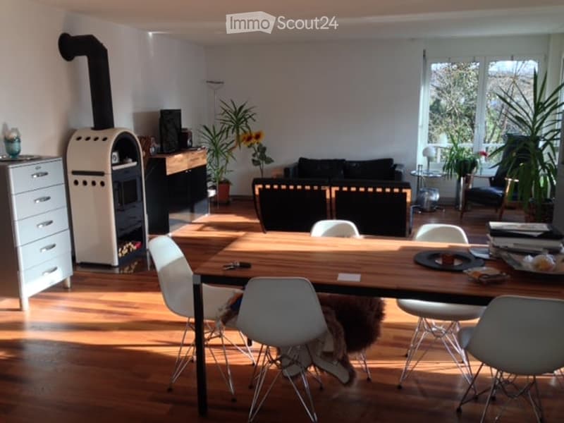 Bright living and dining area with wooden flooring, a large wooden dining table with white chairs, a black sofa, a fireplace, and potted plants.