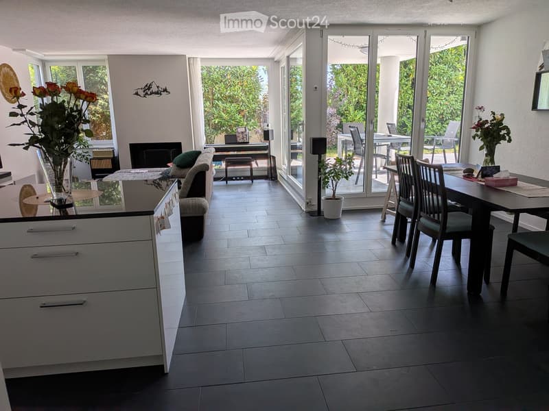 Bright, open living and dining area with large windows and sliding glass doors leading to a patio. Features a sofa, coffee table, dining table with chairs, kitchen cabinetry, and decorative plants.