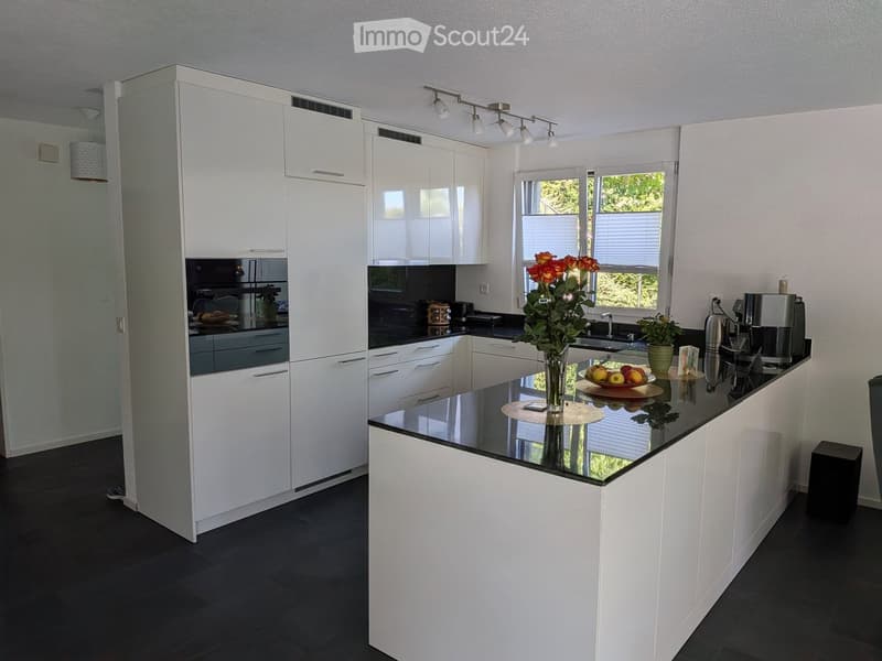 Modern kitchen with white cabinets, black countertops, and stainless steel appliances. Features a central island with a vase of flowers and a bowl of fruit, along with a coffee maker and a potted plant on the counter.