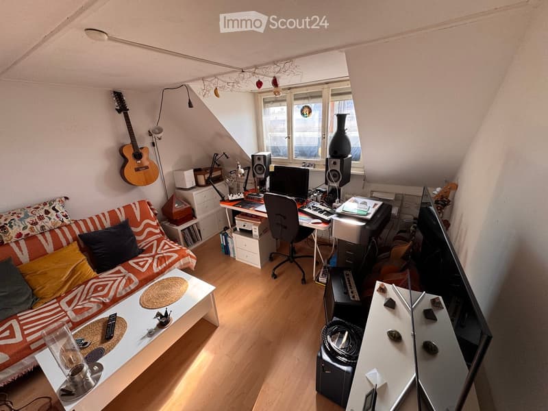 A cozy, multi-functional room with a slanted ceiling, featuring a sofa, coffee table, desk with computer and speakers, and a guitar on the wall.