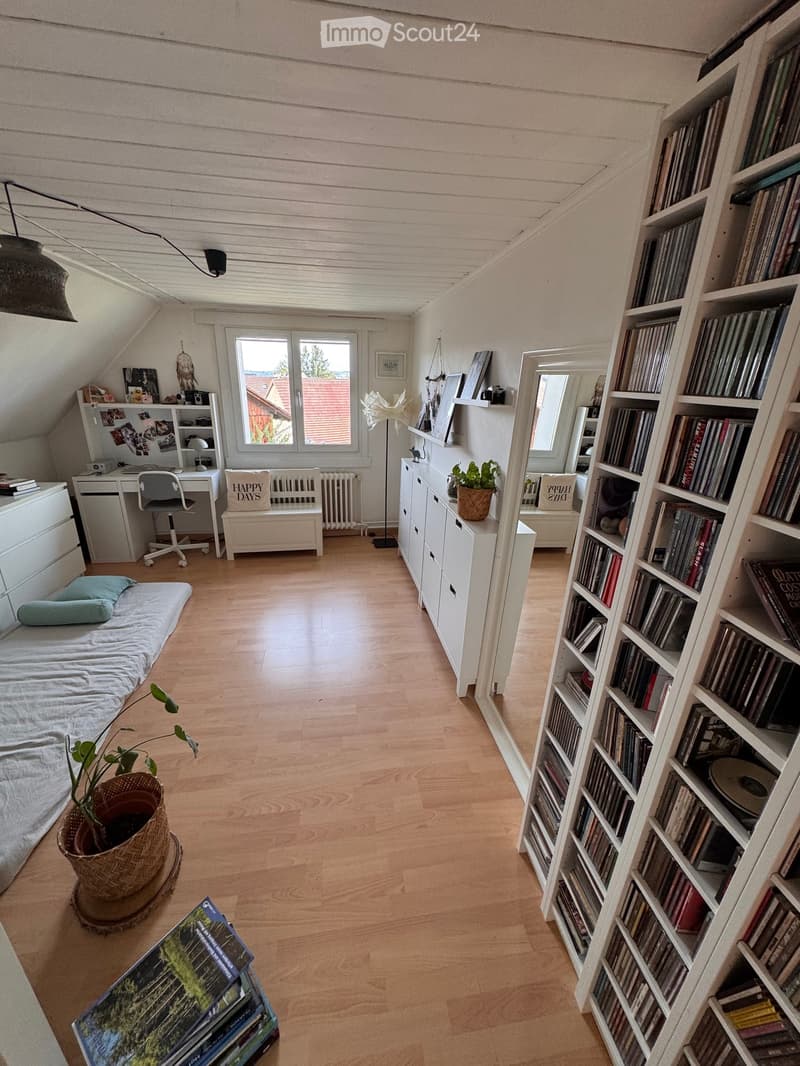 Cozy attic bedroom with wooden floors, slanted ceilings, and large windows. Features a single bed, desk with chair, bookshelf filled with books and CDs, potted plants, and decorative items.