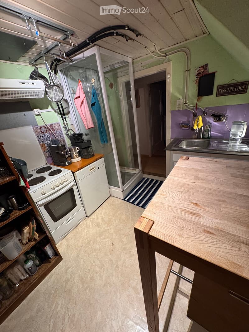 Compact kitchen with white oven, dishwasher, coffee maker, sink, and wooden table. Shower area visible in the background.