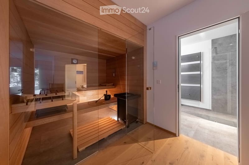 Modern bathroom with wooden sauna bench, black bucket, and black stove, featuring glass walls and wooden flooring.