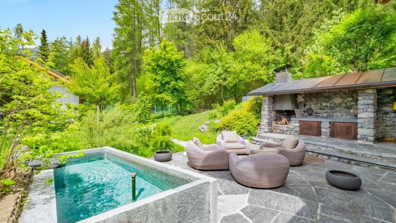 A serene outdoor area with a swimming pool, surrounded by lush greenery and trees. A stone outdoor kitchen with a grill and seating area is visible, along with wicker chairs and a stone pathway.