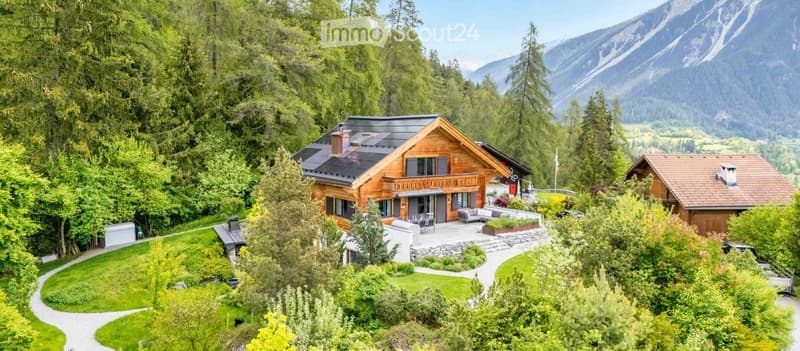 A large, modern wooden house with a balcony, surrounded by lush greenery and mountains in the background.