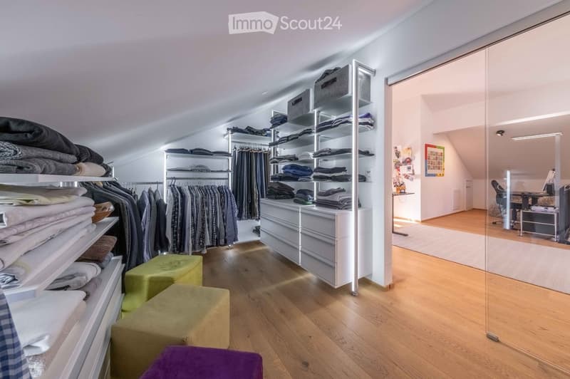 A well-organized walk-in closet with wooden flooring, white walls, and modern built-in storage units. The space includes clothing racks, shelves filled with folded garments, and a small seating area with green and purple stools.