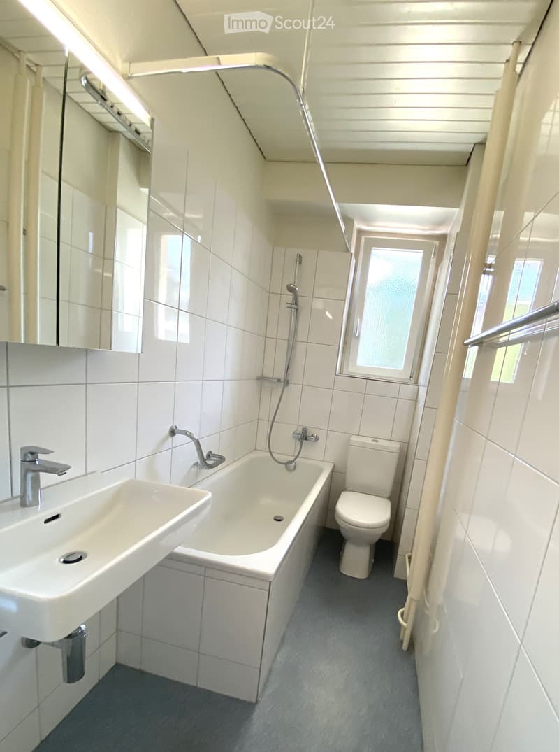 Modern bathroom with white tiles, a sink, bathtub, toilet, and a showerhead. Features a large mirror and a window with frosted glass.