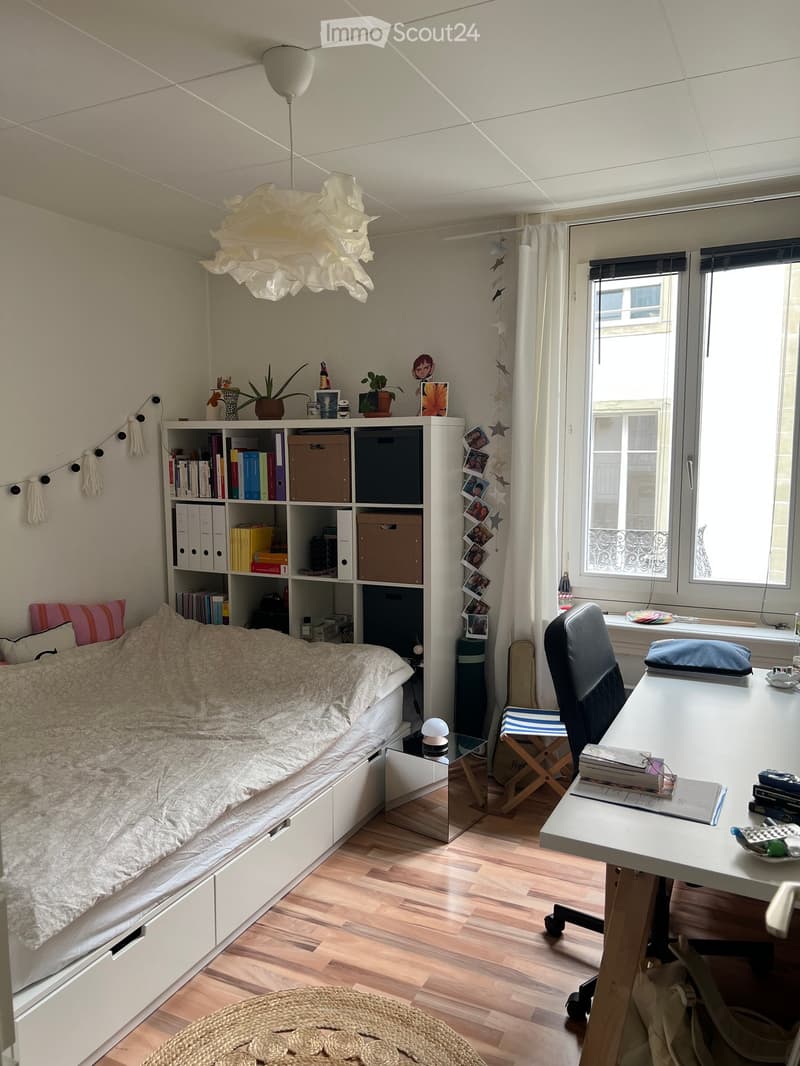 A cozy bedroom with a bed, desk, bookshelf, and a large window with curtains. The room features a hanging lamp, a potted plant, and a small round rug on the wooden floor.