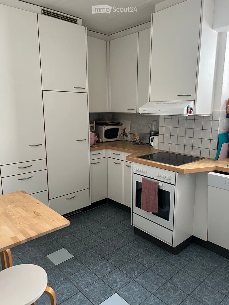 Modern kitchen with white cabinets, wooden countertops, and stainless steel appliances including an oven, microwave, and dishwasher. Features a small wooden table and stool, along with a tiled floor and a range hood.