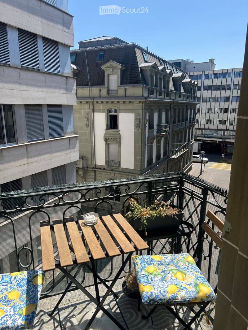 Balcony with a small table, two chairs, and potted plants, overlooking a cityscape with historic buildings.