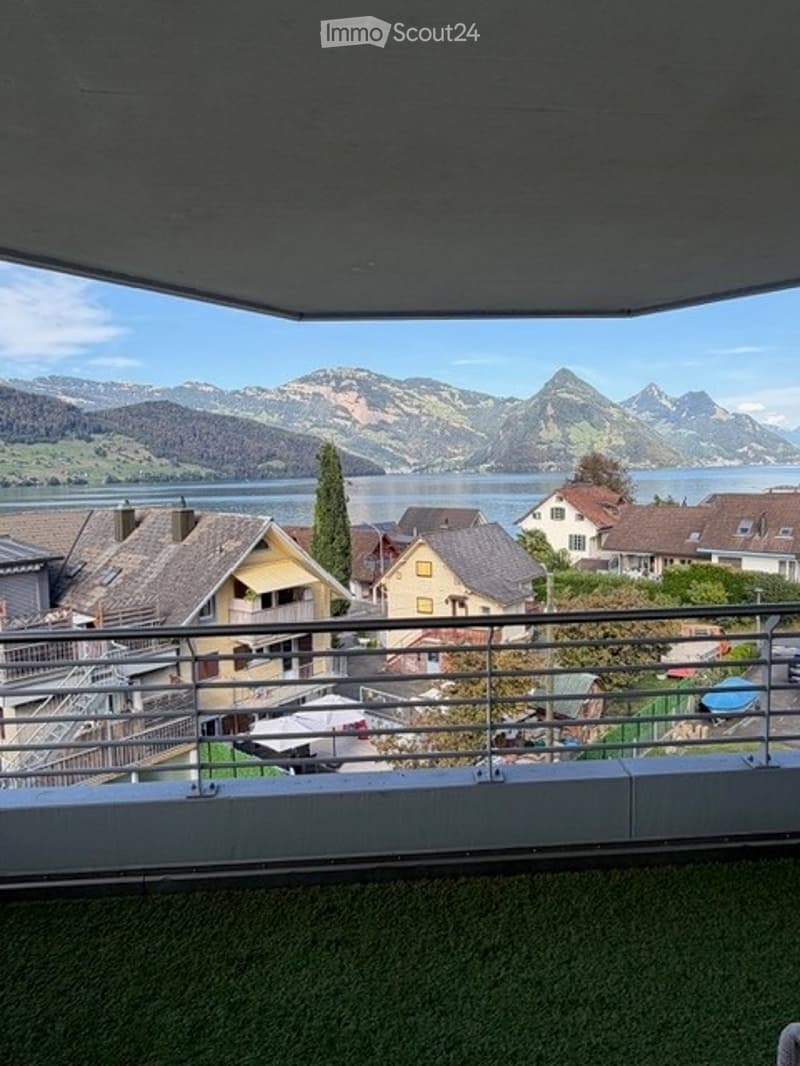 Balcony with metal railing, overlooking a scenic lake and mountain landscape, with houses and trees in the foreground.